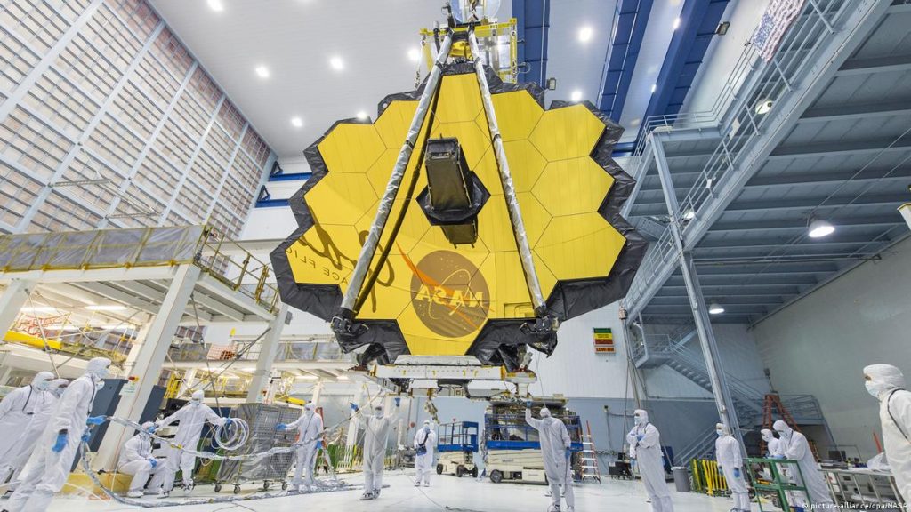 Cientistas da NASA em trajes brancos revisando meticulosamente o Telescópio Espacial James Webb dentro do hangar antes de seu lançamento.